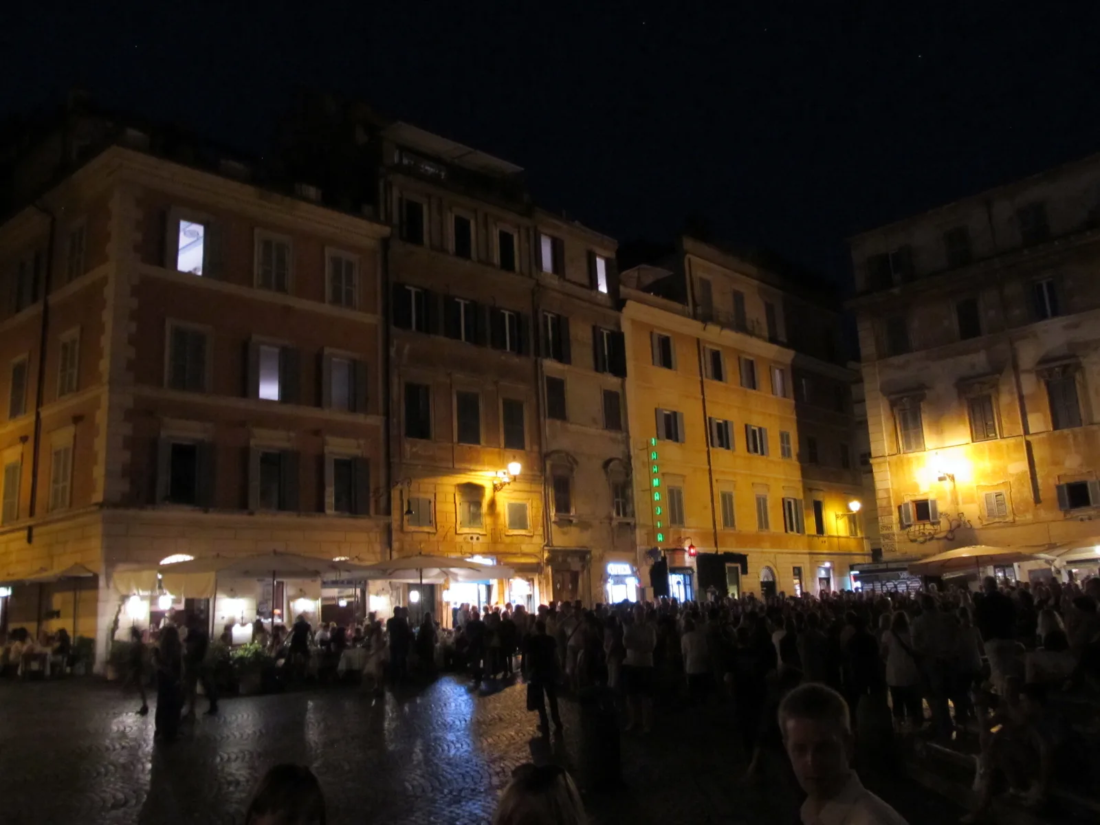 Piazza di Santa Maria in Trastevere a Roma di sera, con la gente seduta intorno alla fontana e ai tavolini dei bar sotto le luci