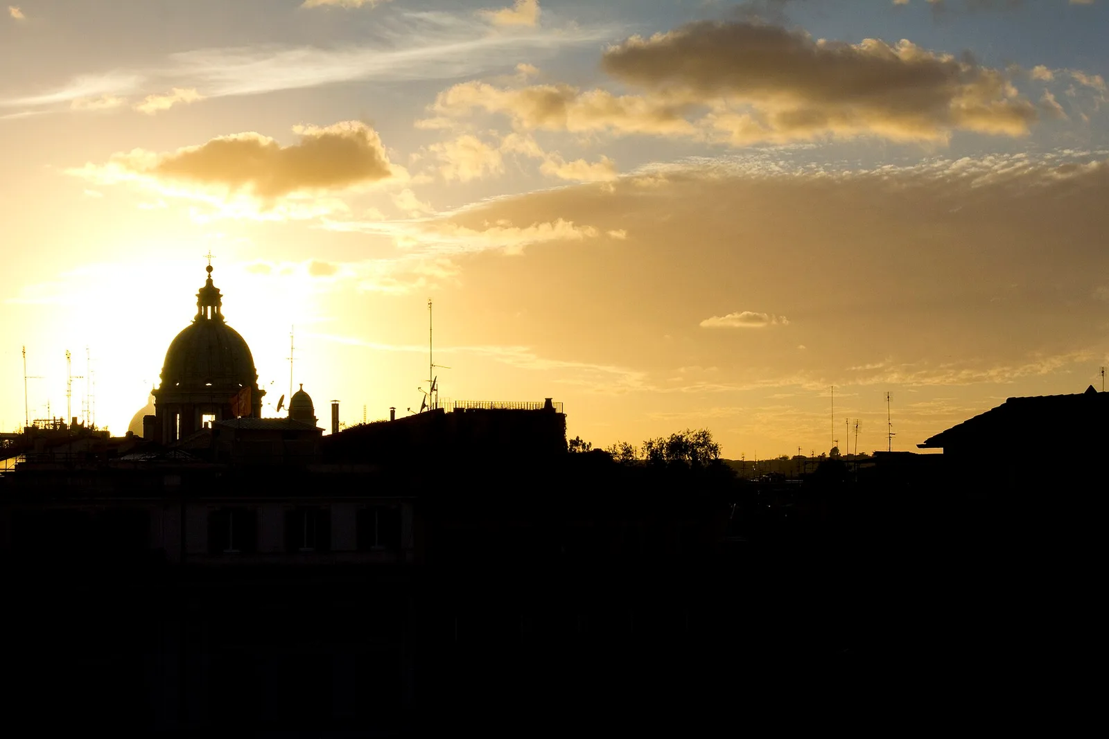 Cupole di Roma in controluce al tramonto, con il sole basso che scende dietro i profili delle chiese e accende il cielo di toni caldi